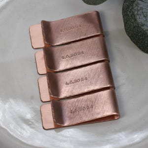 May include: Four copper money clips, each engraved with "4.6.2024". The clips have a brushed finish and a rectangular shape with a curved top. They are arranged on a light gray surface.