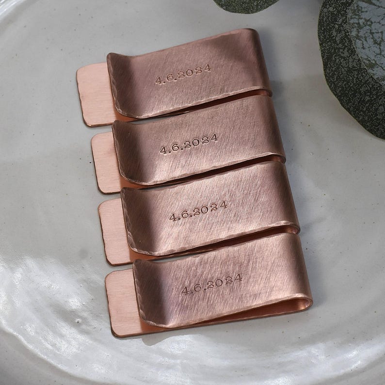 May include: Four copper money clips, each engraved with "4.6.2024". The clips have a brushed finish and a rectangular shape with a curved top. They are arranged on a light gray surface.