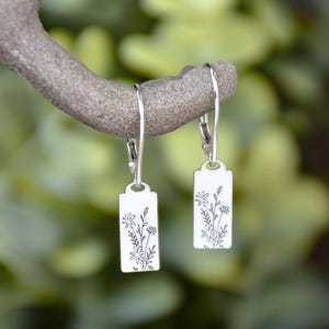 May include: A pair of silver-colored dangle earrings. Each earring features a rectangular tag with a floral design etched in black. The earrings are suspended from a silver hook and are displayed against a blurred green background.