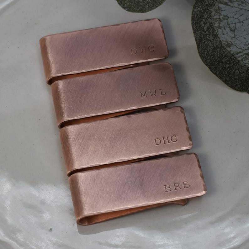 May include: Four copper money clips with hammered texture, each engraved with initials: DWC, MWL, DHC, and BRB. The clips are arranged in a stack, showcasing their warm, metallic color and personalized details. A stylish accessory.