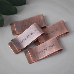 May include: Four copper-colored money clips with the text "Class of 2024" engraved on each. The clips have a curved design and a brushed metal finish. The clips are arranged on a light gray surface.