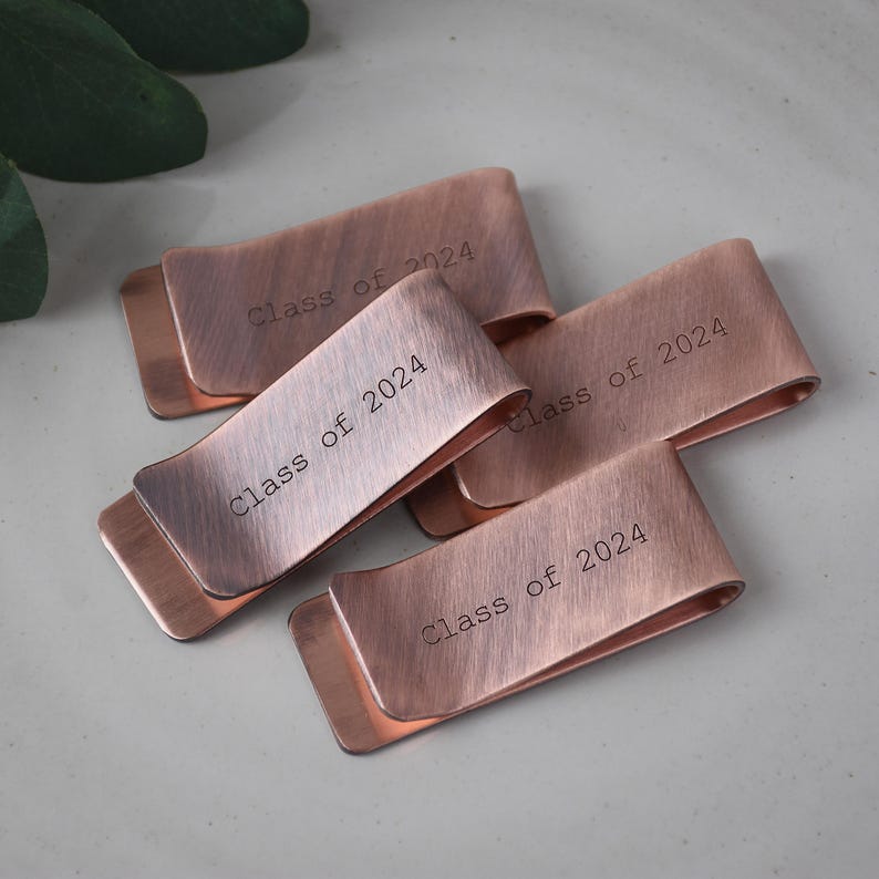 May include: Four copper-colored money clips with the text "Class of 2024" engraved on each. The clips have a curved design and a brushed metal finish. The clips are arranged on a light gray surface.