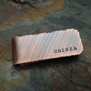 以下が含まれることがあります： ブラシ仕上げの銅製のマネークリップで、"CHINEA"という名前が刻まれています。