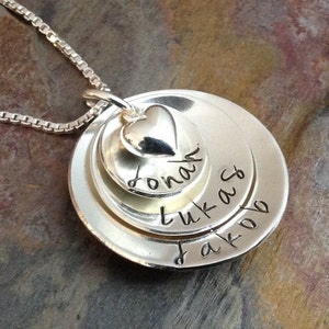 May include: Silver necklace with three layered discs. The top disc has a heart charm and the inscription "Jonah". The middle disc has the inscription "Lukas" and the bottom disc has the inscription "Jakob".