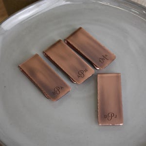 May include: Four copper-colored money clips with a brushed finish are arranged on a gray plate. Three clips have engraved initials, and one clip is blank. The initials are DPA, APM, MA, and DPJ.