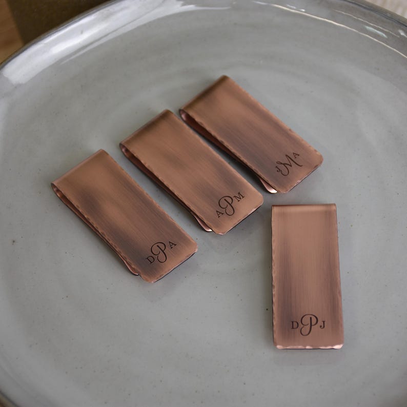 May include: Four copper-colored money clips with a brushed finish are arranged on a gray plate. Three clips have engraved initials, and one clip is blank. The initials are DPA, APM, MA, and DPJ.