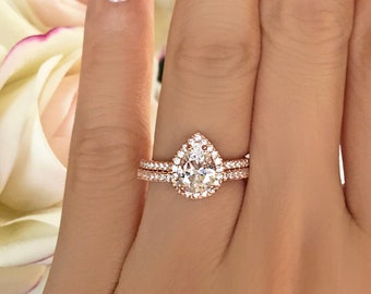 May include: Rose gold engagement ring set with a pear-shaped center stone and a pavé band. The ring is shown on a hand with French-manicured nails, with a bouquet of white and pink roses in the background.