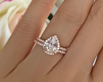 May include: A pear-shaped diamond engagement ring set in rose gold with a halo of smaller diamonds. The ring is worn on a hand with a French manicure.