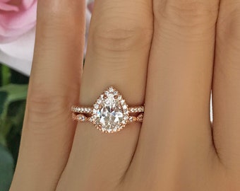 May include: A rose gold pear-shaped engagement ring with a halo setting and a matching band. The ring is on a hand with white french manicured nails.