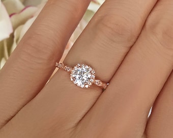 May include: A rose gold engagement ring with a round center stone and a halo of smaller stones. The band is delicate and features a twisted design.