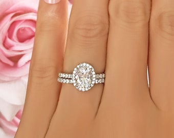 May include: A silver ring set with a large oval-shaped diamond surrounded by smaller diamonds. The ring is on a hand with white French manicured nails.