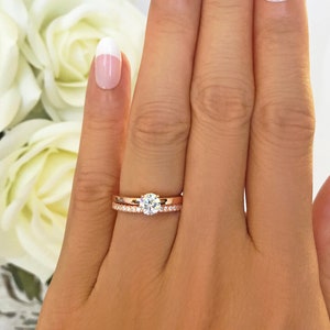 May include: A close-up of a hand wearing a rose gold engagement ring with a round diamond and a matching rose gold wedding band.