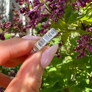 May include: A silver ring with multiple rectangular-cut, clear gemstones. The ring is held in front of a background of purple flowers and green leaves. The gemstones are set closely together, creating a continuous sparkle.