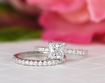 Puede incluir: Un anillo de compromiso de plata con una piedra central de diamante de corte cuadrado y una banda a juego con diamantes más pequeños.
