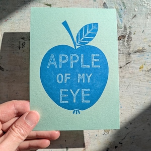 May include: A light blue greeting card with a large blue apple illustration. The apple contains the text "APPLE OF MY EYE" in a white, sans-serif font. The card is held by a hand.