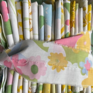 May include: A bundle of folded fabric strips featuring various patterns in shades of yellow, pink, green, and light blue. The fabric pieces are held together by a horizontal band of white cloth printed with a repeating floral design in pastel colors.