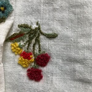 May include: A white linen fabric featuring hand-stitched embroidery of a cluster of berries and leaves. The embroidery includes two yellow berry clusters, two dark red berry clusters, and green stems and leaves, with a partial blue flower.