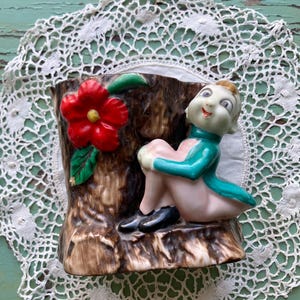 May include: A ceramic wall planter shaped like a tree trunk with a small elf figurine sitting on it. The elf is wearing a teal jacket and has a pink face. A red flower is growing out of the tree trunk.