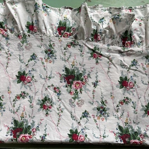 May include: A white fabric bed skirt featuring a repeating floral pattern with red roses, green leaves, and delicate blue accents. The fabric has a gathered ruffle along the top edge, creating a textured, layered appearance across the entire surface.