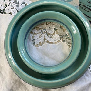May include: Two circular, teal-colored ceramic bowls nested within each other. The bowls feature a smooth, glossy finish with a darker blue-green rim detail. The ceramic material has a consistent, rounded shape with a wide, open center.