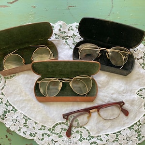 May include: An assortment of vintage eyeglasses and cases. Three pairs of gold-rimmed glasses are displayed in open cases with green velvet lining. A fourth pair of glasses with a red and gold frame rests below. The items are on a white lace doily.