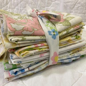 May include: A stack of vintage fabric pieces, featuring floral patterns in pink, yellow, and blue. The fabric is tied with a patterned ribbon, creating a bundle. The fabrics have a variety of floral designs, perfect for quilting or crafting projects.