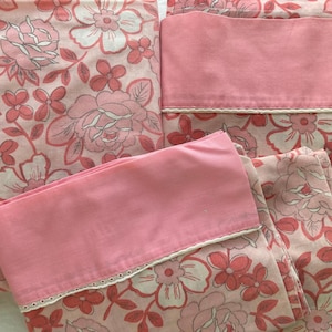 May include: A set of pink floral bed sheets and pillowcases. The sheets feature a pattern of large pink and white flowers with green stems and leaves. The pillowcases are solid pink with a white lace trim.