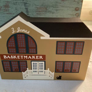May include: A wooden model of a shop with a brown roof and tan walls. The shop has a sign that reads "J. Jones Basketmaker". There are windows with red and blue panes and a white porch with a black cat.