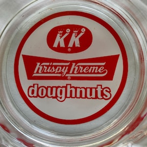 May include: A clear glass dish featuring a circular red and white logo for Krispy Kreme Doughnuts. The design includes the letters KK inside an oval, the brand name Krispy Kreme in a stylized script, and the word doughnuts in bold red letters.