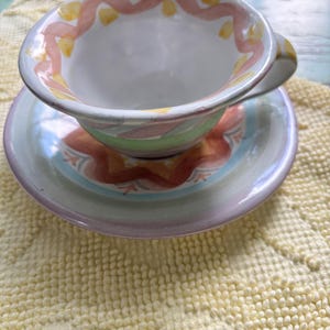 May include: A ceramic teacup and saucer set featuring a hand-painted design. The cup has a white interior with a pink wavy line and yellow dots along the rim. The exterior and saucer display horizontal bands of green, pink, and orange patterns.