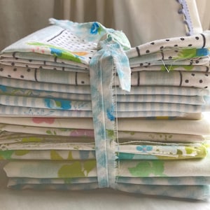 May include: A stack of folded vintage fabric pieces, tied with a light blue ribbon. The fabrics display various patterns, including polka dots, floral designs, and stripes, in shades of blue, green, and white. The bundle is neatly arranged, suggesting a collection of textiles.