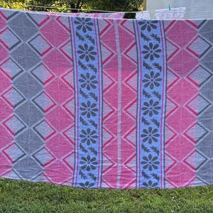 May include: A rectangular textile featuring a repeating pattern of vertical stripes. The design alternates between bands of pink and gray diamonds and light blue columns decorated with dark gray floral motifs and small leaf-like shapes.