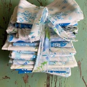 May include: A stack of folded fabric squares tied with a matching ribbon. The fabric features a blue floral pattern on a white background. The ribbon is a mix of blue and green floral patterns.