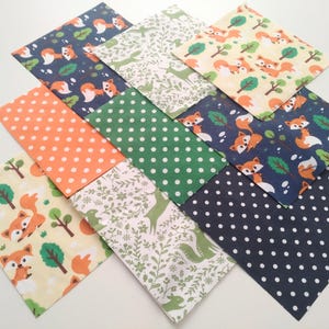 May include: A collection of patterned fabric squares featuring foxes, trees, deer, and polka dots. The colour palette includes orange, green, navy, and cream. The fabrics are arranged in an overlapping design, creating a visually interesting display.