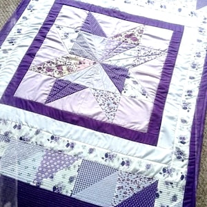 May include: A purple and white patchwork quilt with a star design in the center. The quilt is made of various fabrics with floral patterns and different shades of purple.