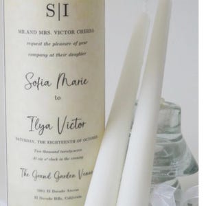 May include: A tall, cylindrical candle with printed text, next to two white taper candles. The candle has a cream-colored label with the initials "S|I" and wedding invitation details. The taper candles are slender and white, suitable for a wedding ceremony.