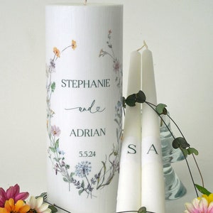 May include: Two white taper candles with the initials "S" and "A" printed on them, and a larger white pillar candle with a floral wreath design and the text "STEPHANIE and ADRIAN 5.5.24".