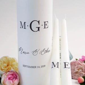 May include: A white pillar candle and two taper candles with black lettering. The pillar candle has the initials "M.G.E", the names "Marie & Ethan", and the date "September 14, 2028". The taper candles have the initials "M E".