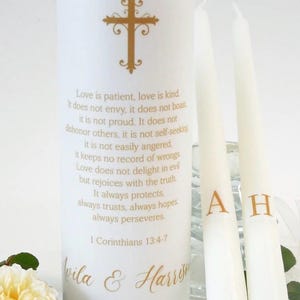May include: A large white pillar candle featuring a gold cross, the text 1 Corinthians 13:4-7, the names Vila & Harrison, and the date April 12, 2028. Two thinner white taper candles stand beside it, each marked with a single gold letter, A and H.