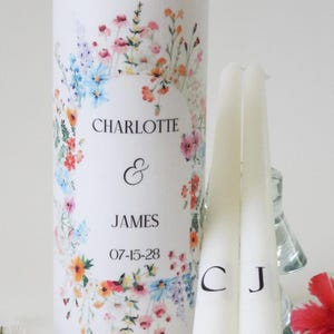May include: A large white pillar candle decorated with a colorful wildflower wreath, featuring the text CHARLOTTE & JAMES and the date 07-15-28. Beside it are two slender white taper candles, each marked with a single black letter, C and J.