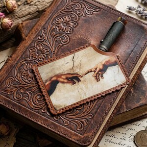 Könnte beinhalten: Zwei braune Lederjournale mit floralen Prägungen. Ein Stift liegt obenauf und ein Lesezeichen aus Leder mit einem Renaissance-Gemälde von sich berührenden Händen. Getrocknete Blumen und Münzen sind um die Journale verstreut.
