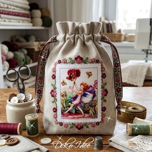 Könnte beinhalten: Ein beigefarbener Leinenbeutel mit Kordelzug, der ein zentrales rechteckiges Feld mit dem Aufdruck einer Fee mit einer großen roten Blume zeigt, umgeben von gestickten roten und rosa Blüten und dekorativen Bändern an den Seiten.