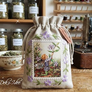 Könnte beinhalten: Ein beigefarbener Leinenbeutel mit Kordelzug und einer zentralen rechteckigen Illustration einer Fee zwischen violetten Blumen. Der Beutel ist mit gestickten violetten Blumen um das Bild herum verziert und hat ein Holzschild mit der Aufschrift Handgemacht.