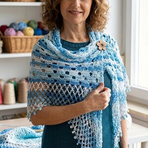 May include: A blue and white crocheted shawl with a floral wooden pin. The shawl is draped over the shoulders. The background includes baskets of yarn and spools of thread, suggesting a craft or handmade item.