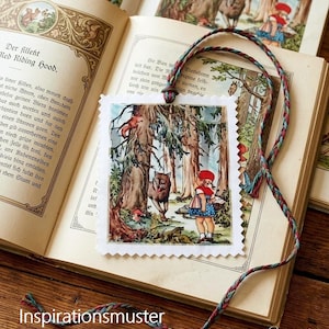 Könnte beinhalten: Ein rechteckiges Lesezeichen aus Papier mit gezackten Rändern, das eine farbenfrohe Illustration eines Mädchens mit roter Kapuze und eines Wolfes im Wald zeigt. Das Lesezeichen hat eine geflochtene, mehrfarbige Kordel und liegt auf einem aufgeschlagenen alten Buch.