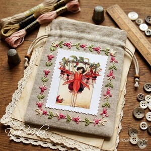 Könnte beinhalten: Ein beigefarbener Leinenbeutel mit Kordelzug, der in der Mitte einen rechteckigen Aufnäher mit einer gedruckten Illustration einer Fee in rotem Blumenoutfit zeigt, umrandet von einer Stickerei aus rosa und grünen Blumen mit Holzperlen.