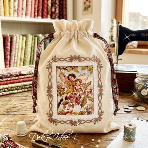 May include: A cream-colored fabric drawstring bag featuring a central rectangular illustration of two winged fairies sitting among pink blossoms. The bag has a gathered top and decorative floral patterned ribbon drawstrings on both sides.