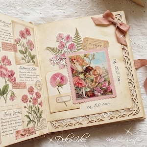 May include: An open scrapbook with cream-colored pages featuring botanical illustrations of pink flowers, a vintage ticket, and a central rectangular card depicting two fairies. The page includes handwritten text and measures approximately 10.0 cm by 8.0 cm.