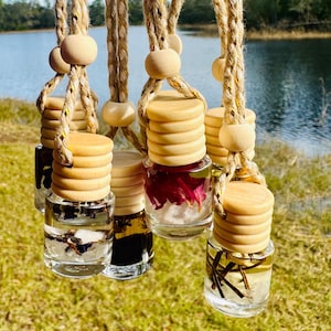 May include: Clear glass car air fresheners with wooden lids and hanging rope. The bottles contain various dried botanicals and liquids. The background shows a lake and trees.