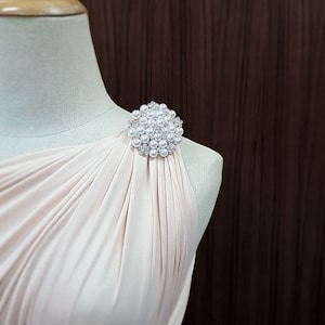 K&ouml;nnte beinhalten: Hellrosa Kleid mit einer Perlen- und Strassbrosche. Die Brosche ist rund und hat mehrere kleine Perlen und klare Strasssteine. Das Kleid ist drapiert und hat ein One-Shoulder-Design.